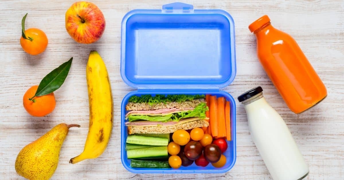 pieces of fruit beside a lunch box full of food and bottles of juice and milk on the right